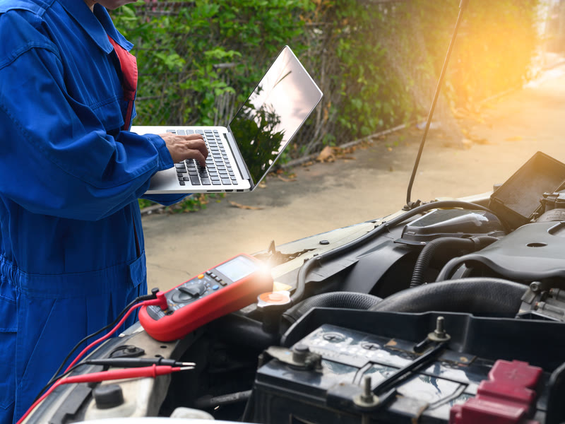 Mechanic in blue coveralls testing a car battery with a multimeter and laptop