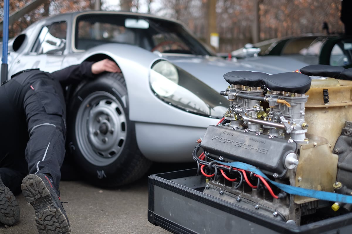 Mechanic working under a silver classic sports car beside its engine